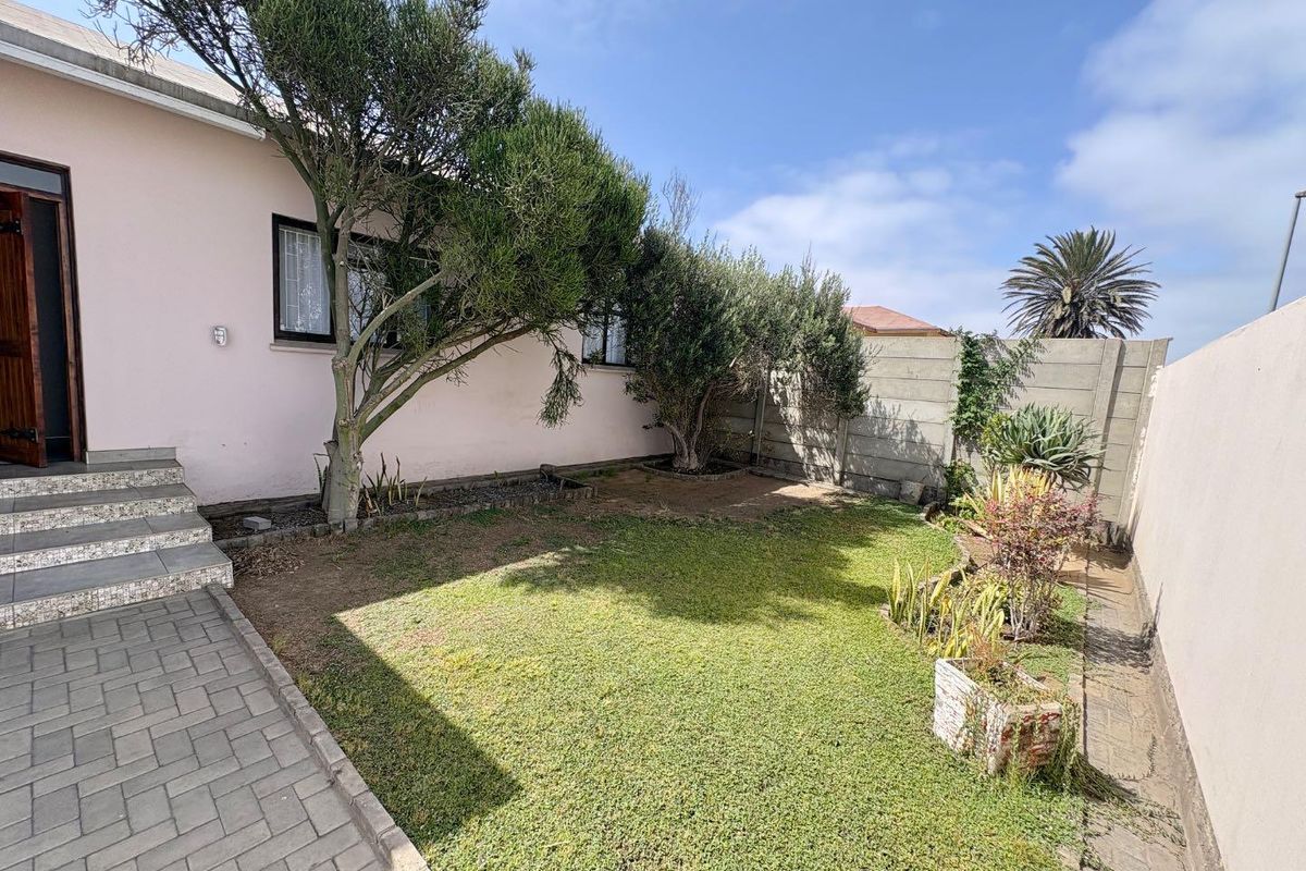 A private rear garden featuring a paved walkway, tiled steps leading to the house, established trees, and a concrete boundary wall.