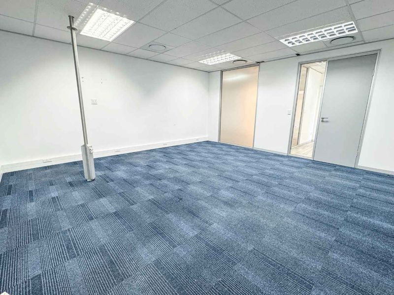 A clean, vacant office space featuring blue carpet tiles, a suspended ceiling with recessed lighting, and a functional power pole for workstation connectivity.