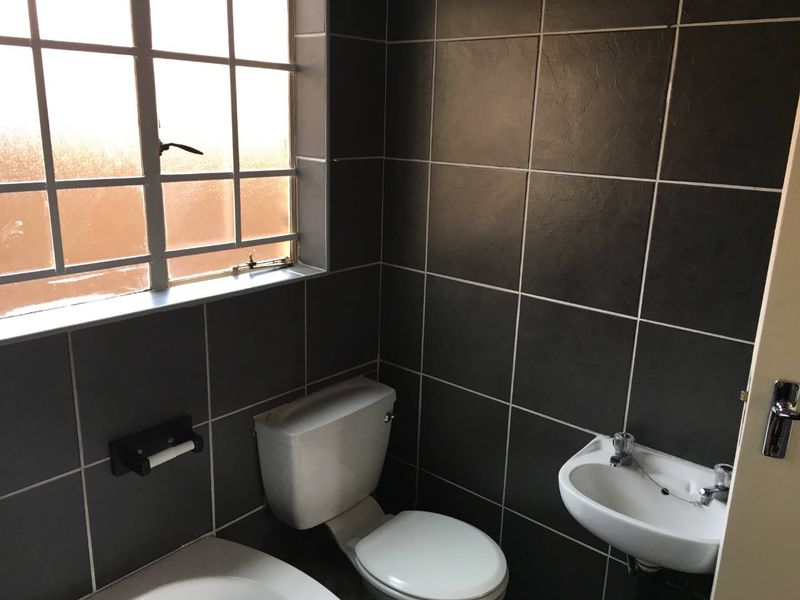 A compact bathroom featuring dark wall tiles, a white toilet, and a wall-mounted basin with a window providing natural light.