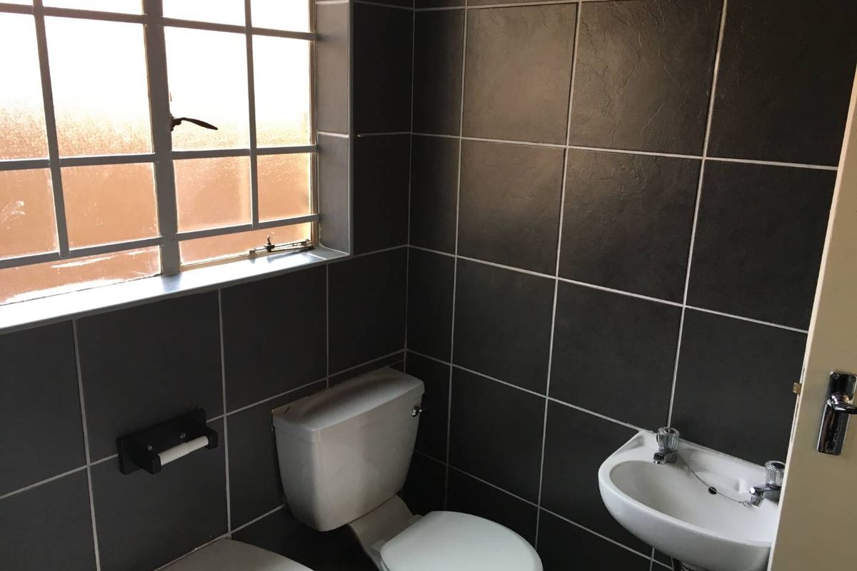 A compact bathroom featuring dark wall tiles, a white toilet, and a wall-mounted basin with a window providing natural light.