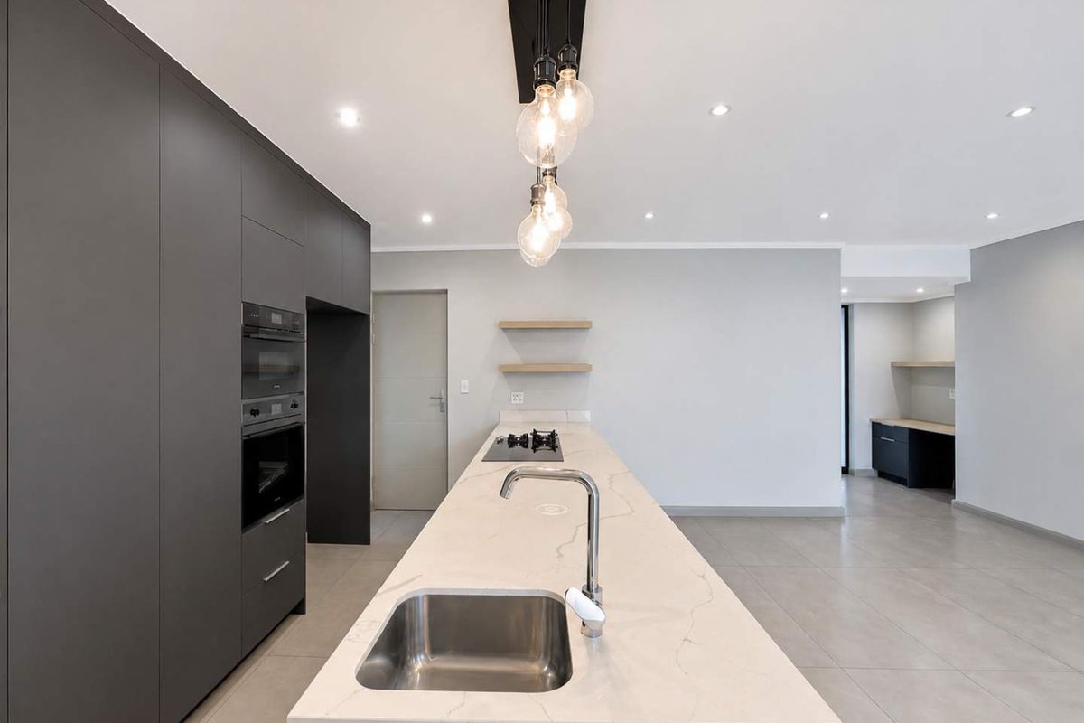 Modern kitchen featuring a large stone-topped island with an undermount sink, built-in appliances, sleek cabinetry, and minimalist floating shelves.