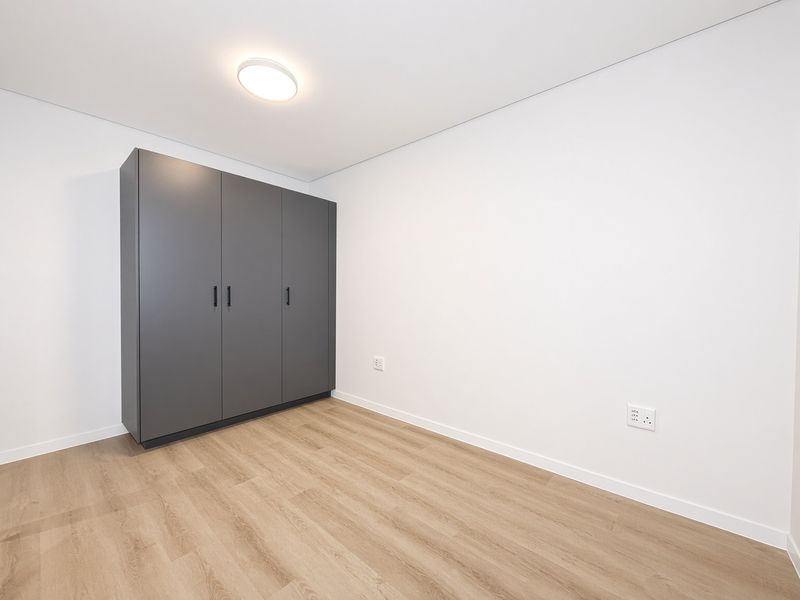 A bright, empty bedroom featuring modern laminate flooring, crisp white walls, and a sleek, dark grey built-in wardrobe.