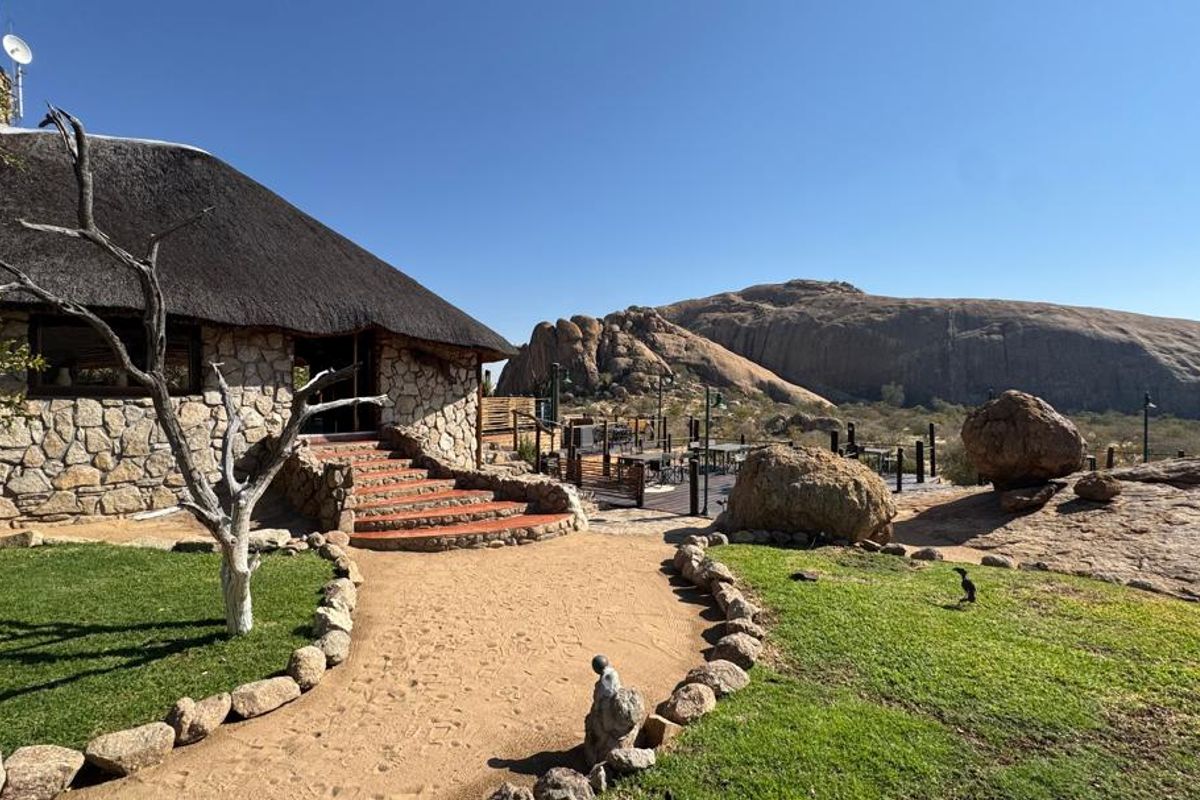 A scenic view of a lodge exterior featuring traditional thatched roofing, stone masonry, and a landscaped garden set against a dramatic rocky mountain backdrop.