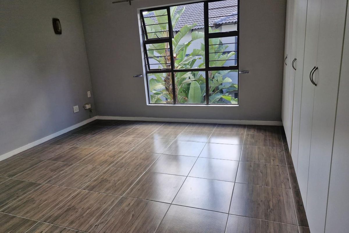 A bright, empty bedroom featuring wood-look tiled flooring, built-in cupboards, and a large window overlooking greenery.