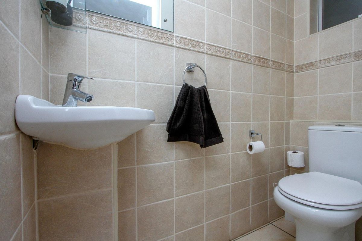 A compact guest bathroom featuring neutral ceramic wall tiling, a white pedestal basin with chrome mixer tap, and a standard toilet.
