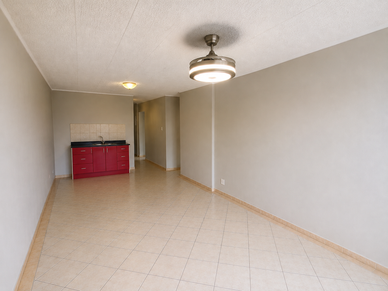 An open-plan living area featuring tiled flooring, a ceiling fan, and a compact kitchenette with red cabinetry and a sink.