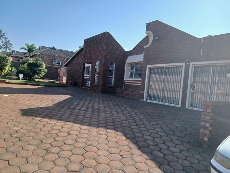 Front exterior view of a brick house featuring a large paved driveway, sliding glass doors with security gates, and established garden surroundings.