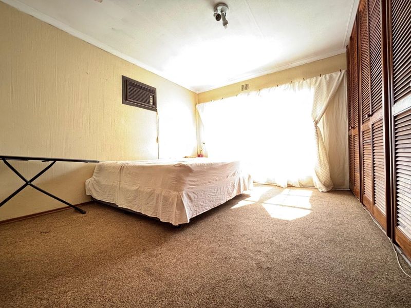 A bedroom featuring built-in wooden louvered cupboards, carpeted flooring, and a wall-mounted air conditioning unit, offering a functional space with potential for modernization.