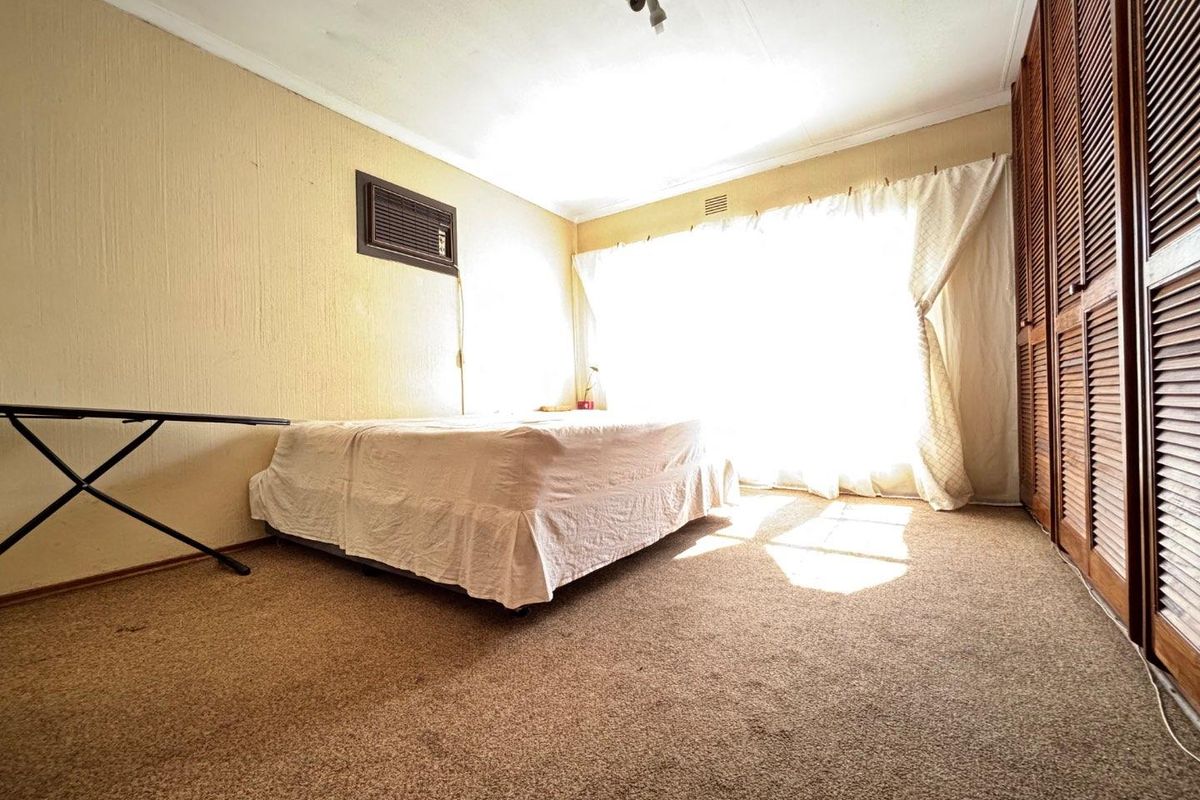A bedroom featuring built-in wooden louvered cupboards, carpeted flooring, and a wall-mounted air conditioning unit, offering a functional space with potential for modernization.