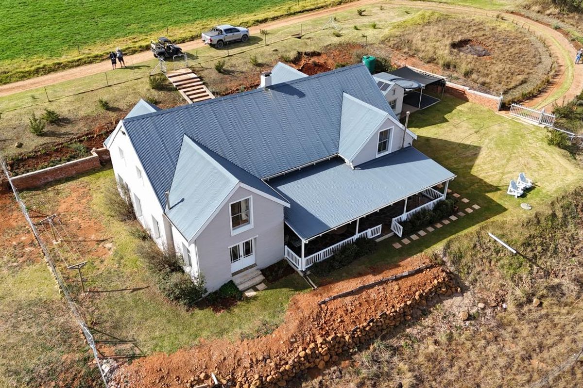 Aerial view of a modern farmhouse with a metal roof, wrap-around veranda, and expansive rural grounds.