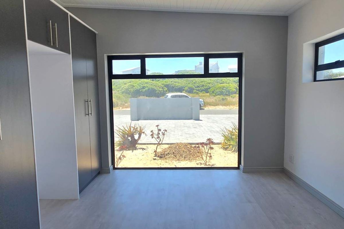 A bright, modern bedroom featuring built-in dark cabinetry, light wood-look flooring, and large windows providing natural light and exterior views.
