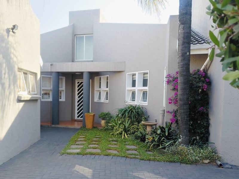Front facade of a townhouse featuring a paved driveway, landscaped garden area, and a covered entrance with security gate and structural columns.