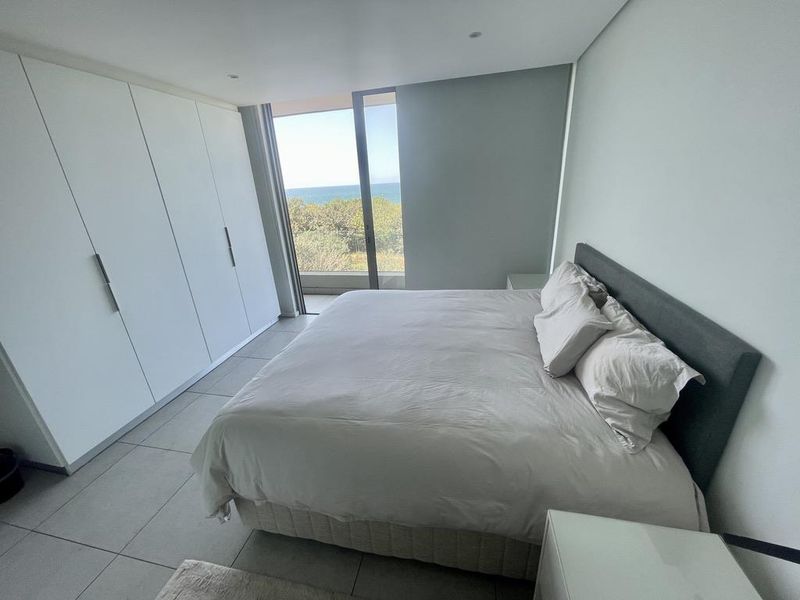 Modern bedroom featuring built-in floor-to-ceiling wardrobes, tiled flooring, and large sliding glass doors opening to a balcony with scenic coastal views.