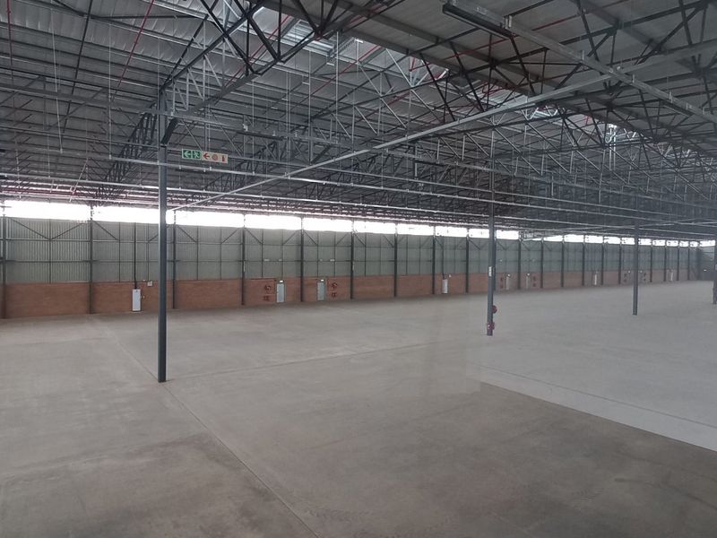A large, open-plan industrial warehouse space featuring high ceilings with exposed steel trusses, concrete flooring, and brick wall construction.
