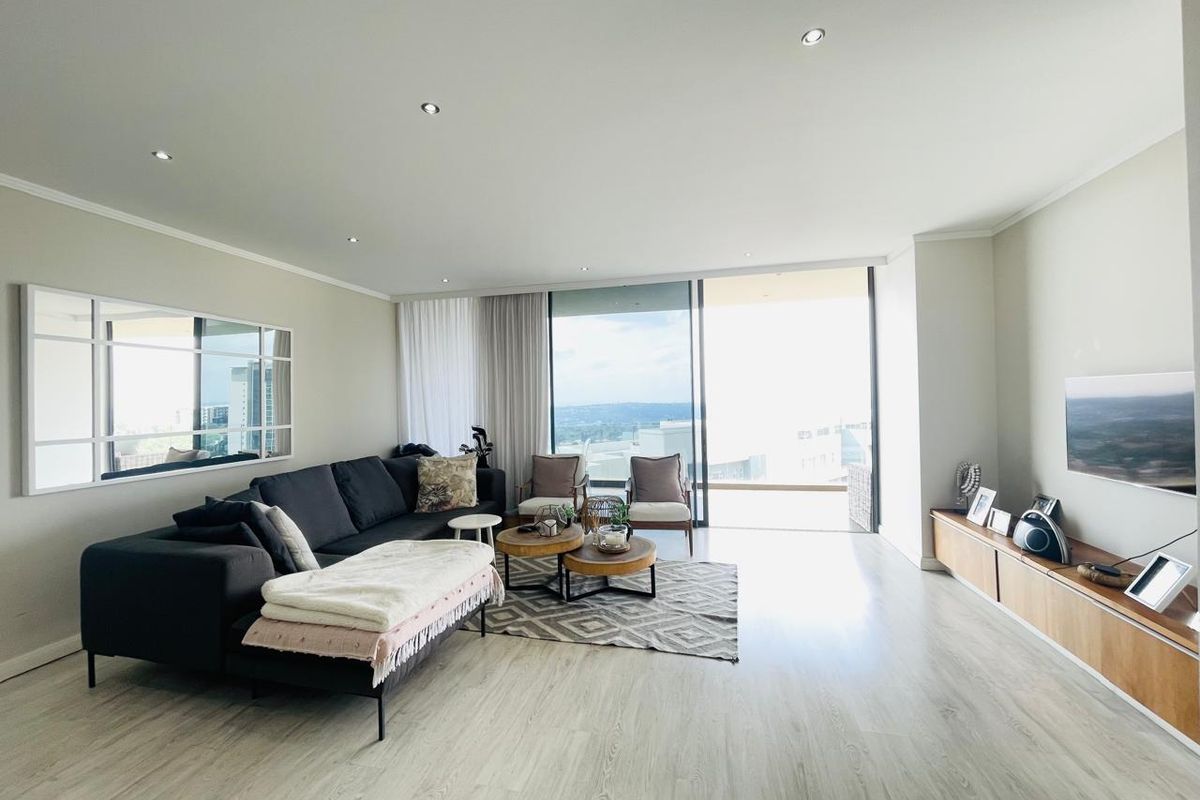 Spacious open-plan living area featuring light wood-look flooring, recessed downlighting, and large sliding glass doors leading to a balcony with expansive views.