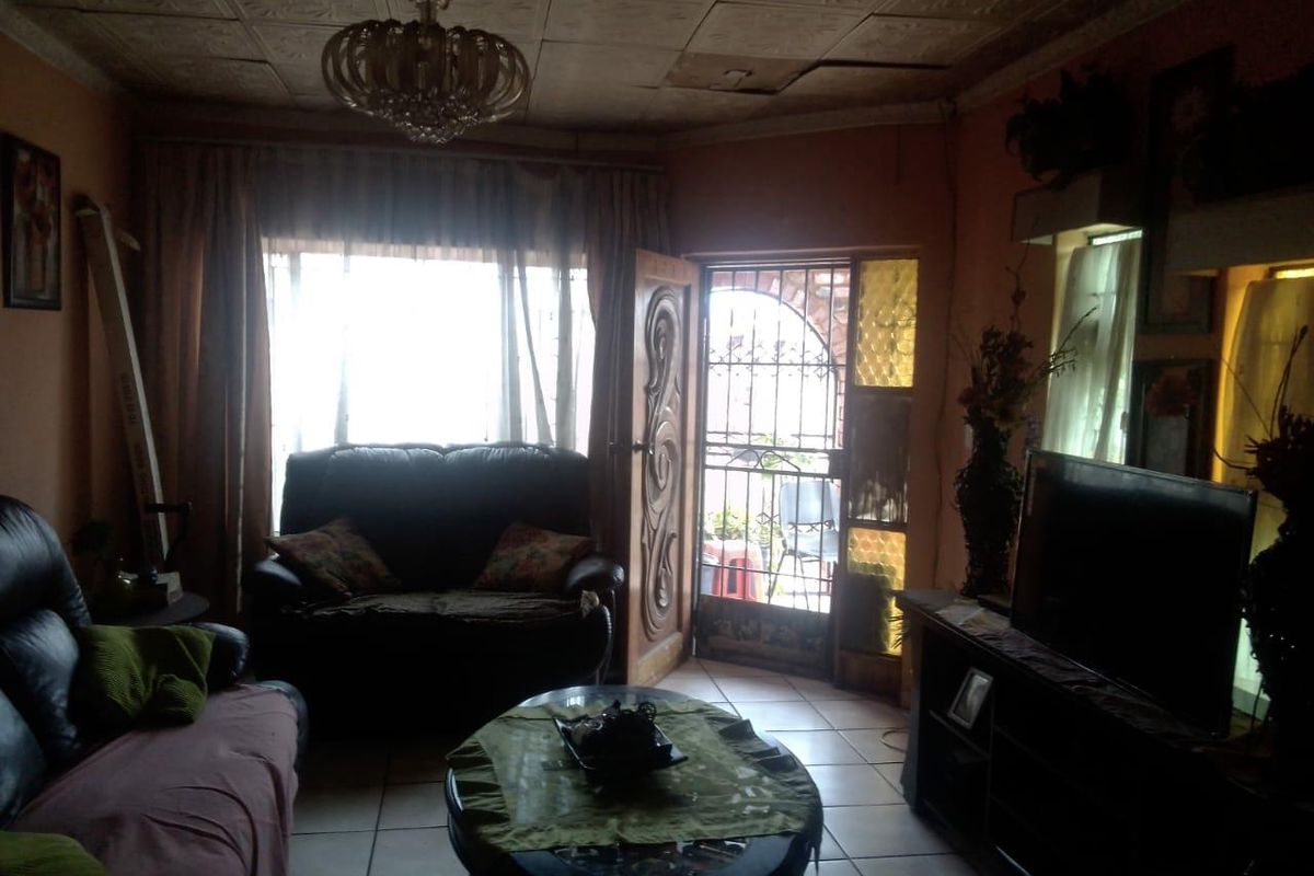 A dimly lit living room featuring tiled flooring, a decorative ceiling, and a security gate entrance, showing signs of significant wear and maintenance needs.