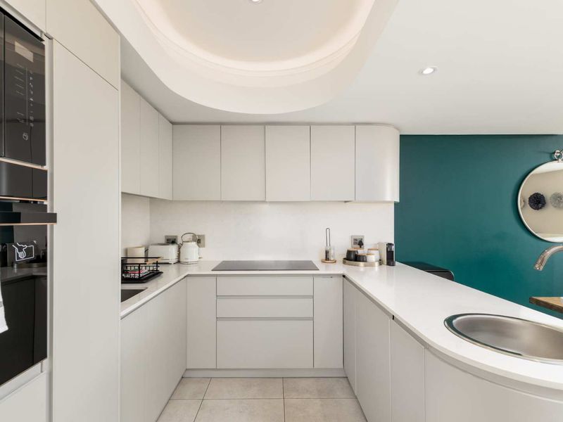 Modern U-shaped kitchen featuring sleek handleless cabinetry, stone countertops, integrated appliances, and elegant cove lighting, complemented by a striking teal accent wall.