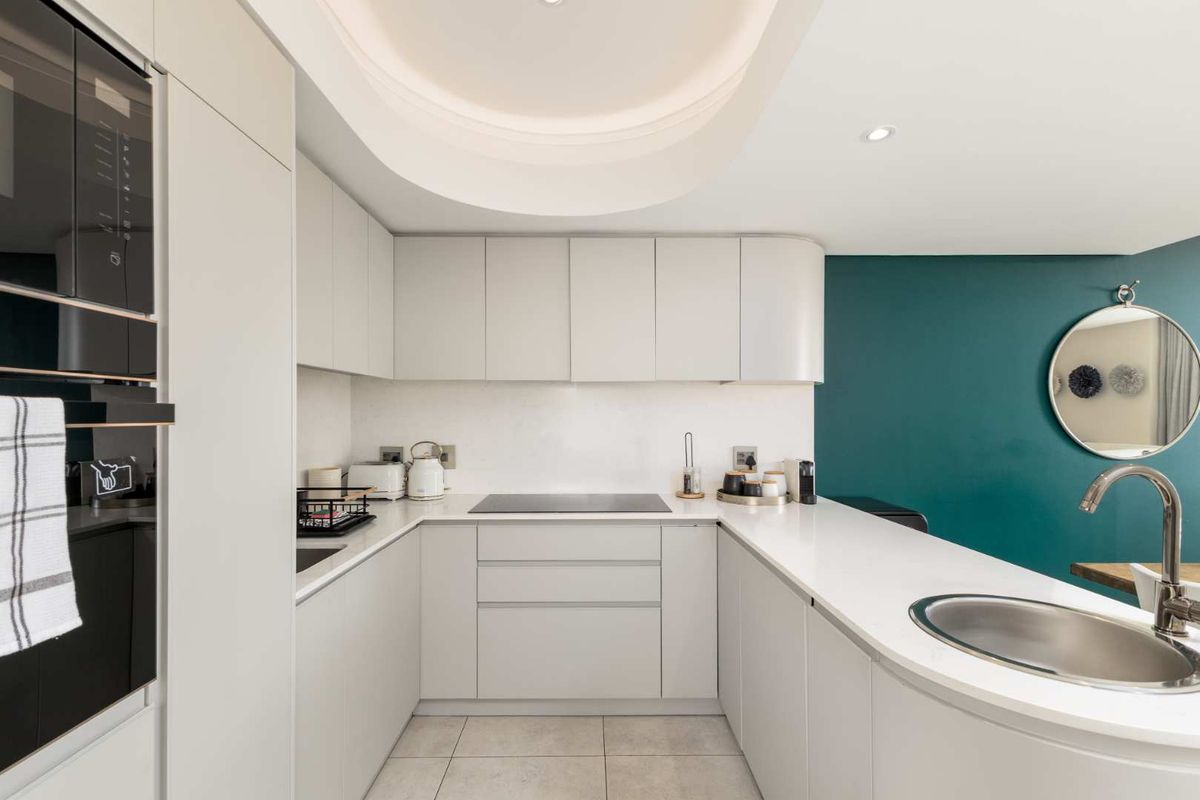Modern U-shaped kitchen featuring sleek handleless cabinetry, stone countertops, integrated appliances, and elegant cove lighting, complemented by a striking teal accent wall.