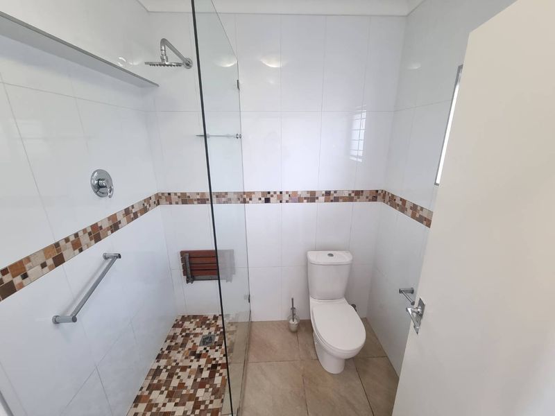 A clean, functional bathroom featuring a walk-in shower with a glass screen, grab rail, fold-down seat, and neutral tiled finishes throughout.