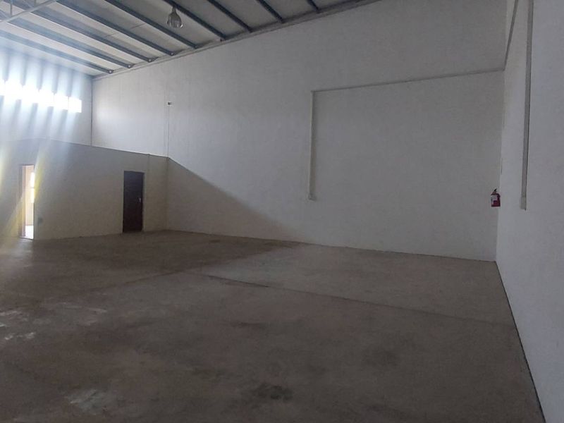 Spacious industrial warehouse unit featuring high ceilings with exposed steel trusses, concrete flooring, and a large roller shutter door for easy access.