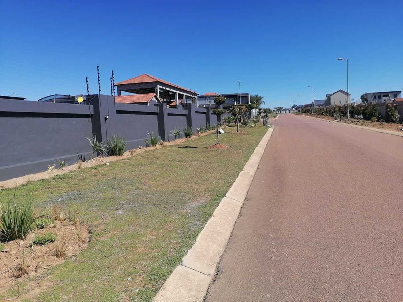 A clear street view of a residential area featuring a paved road, concrete curbing, and a secure perimeter wall with electric fencing.