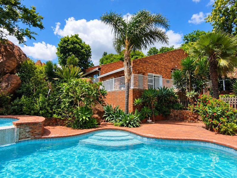 A well-maintained outdoor pool area featuring a swimming pool, raised spa, and brick paving, set against a lush garden and brick house facade.