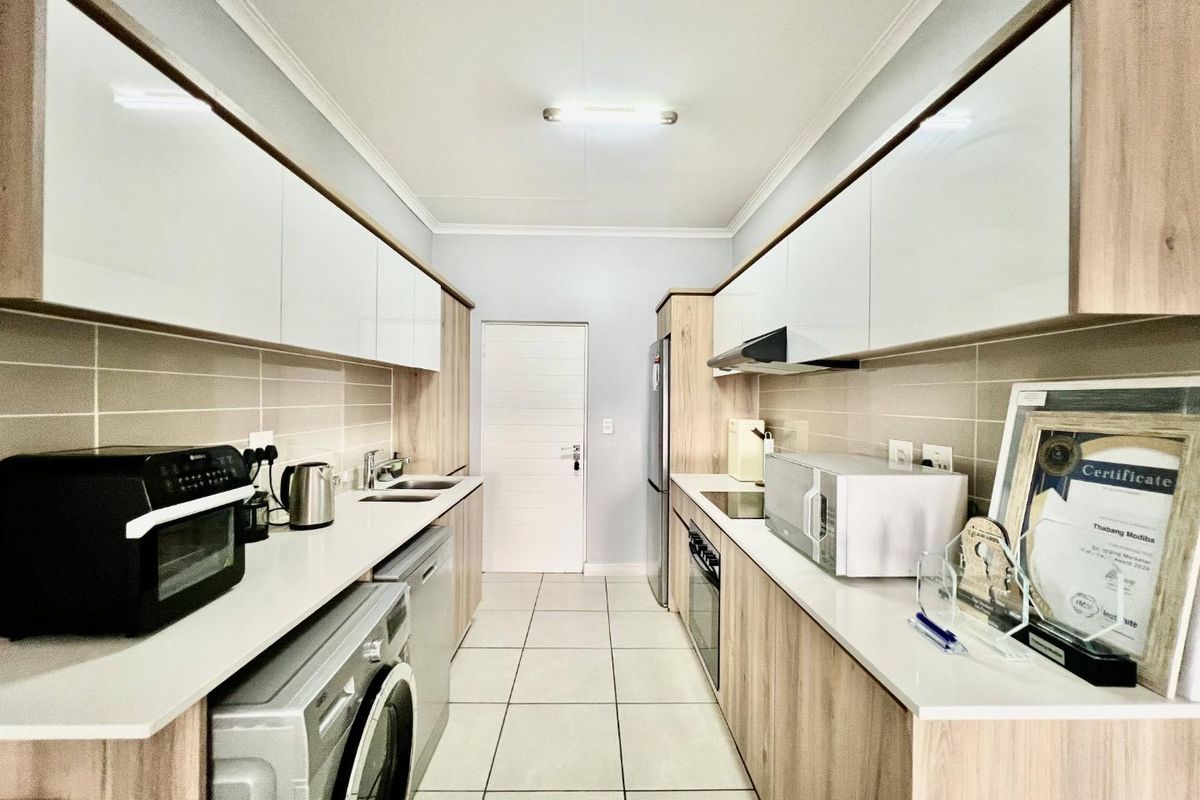 Modern galley-style kitchen featuring sleek white and wood-grain cabinetry, stone countertops, integrated appliances, and neutral tiled flooring.