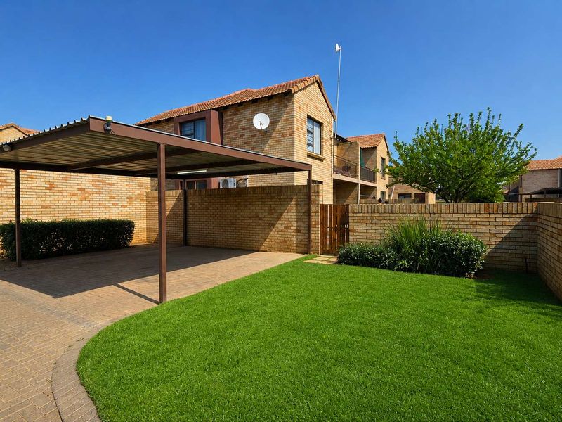 A well-maintained townhouse exterior featuring a paved driveway, a covered carport, and a neat front garden with a brick boundary wall.