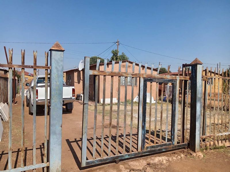 Front view of a residential property featuring a metal security gate, perimeter fencing, and a driveway leading to the main house structure.