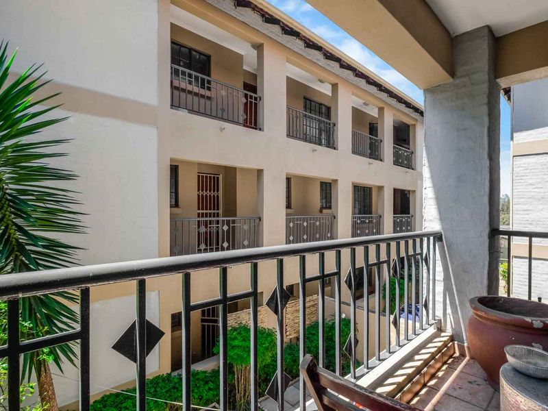 A covered balcony featuring tiled flooring and a metal balustrade, overlooking the apartment complex courtyard.