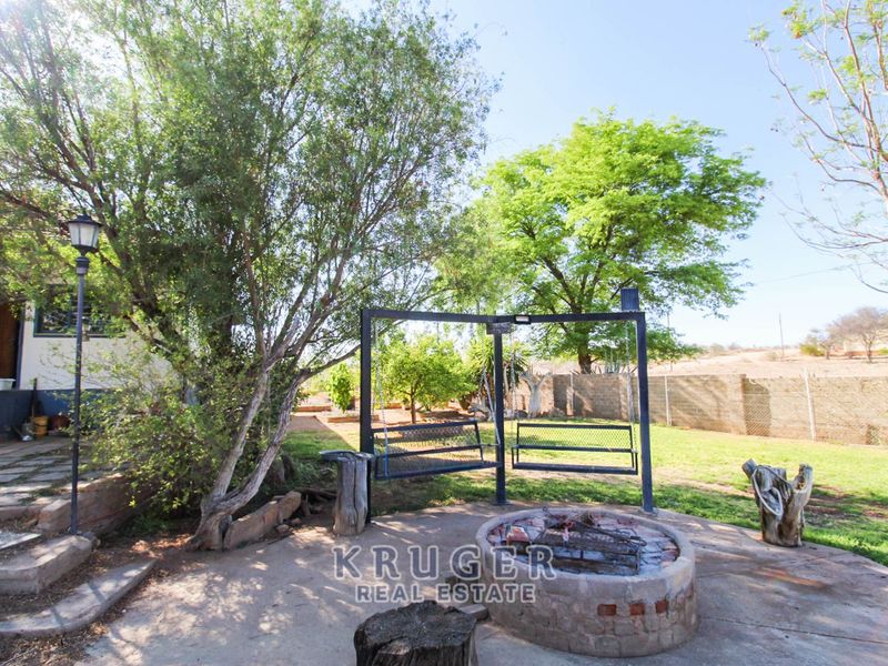 A spacious garden area featuring a built-in brick fire pit, a metal swing set, and a paved patio space surrounded by established trees.