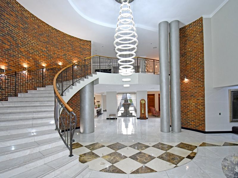 Grand entrance hall featuring a sweeping curved staircase, marble flooring with inlay detail, exposed brick walls, and a striking modern chandelier.