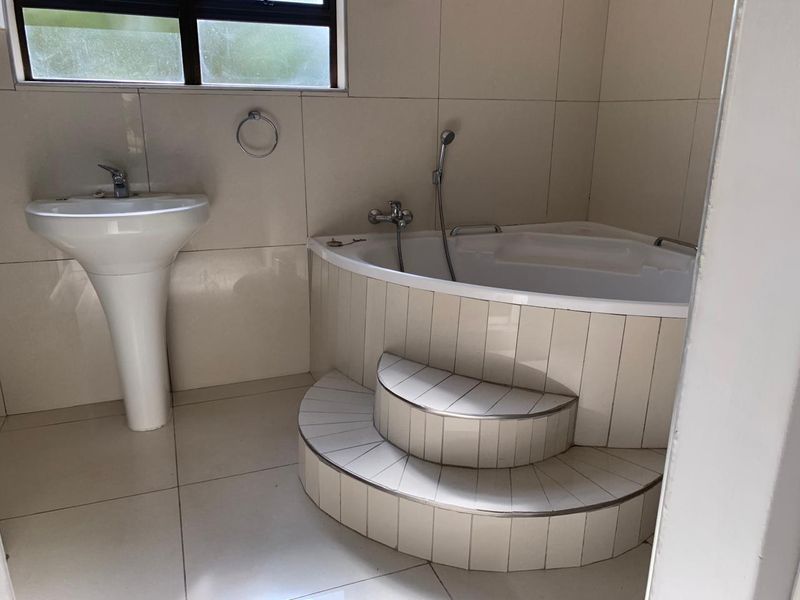 A functional bathroom featuring a corner bathtub with tiled steps, a pedestal basin, and neutral wall tiling.