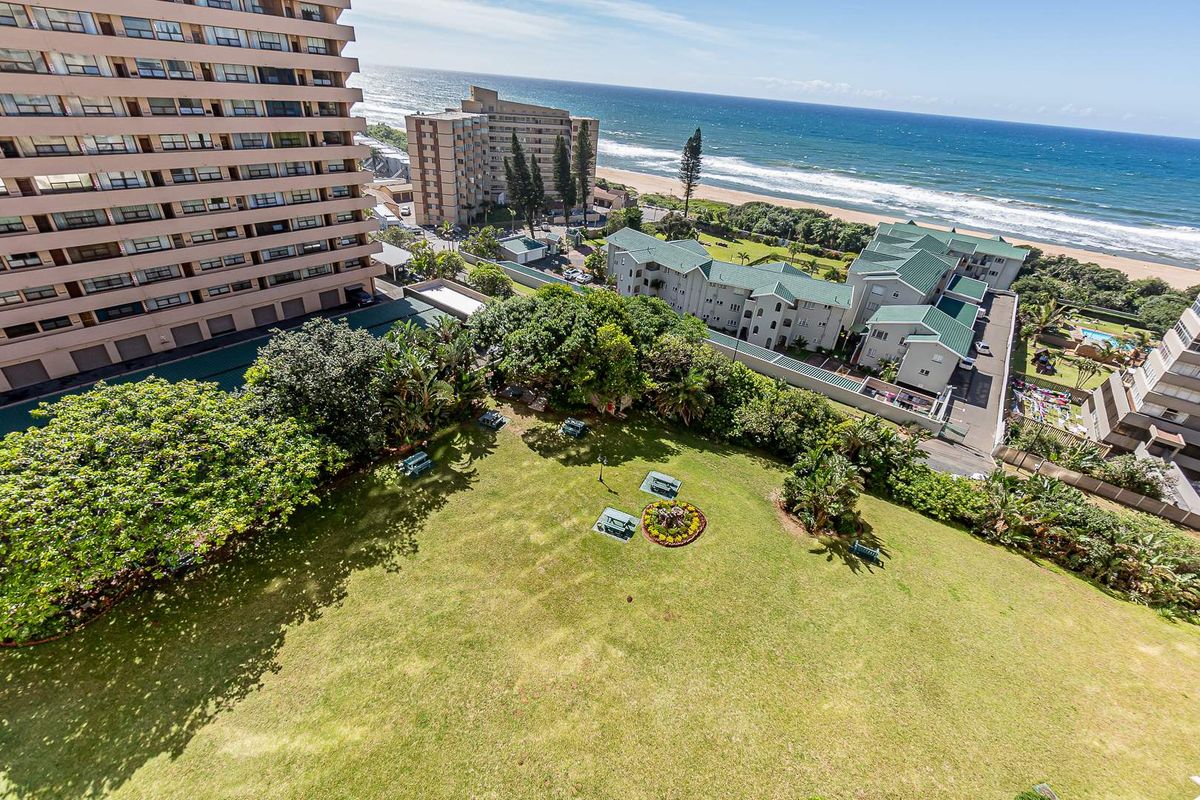 Elevated aerial view overlooking a landscaped communal garden area with mature trees and ocean views in the background.