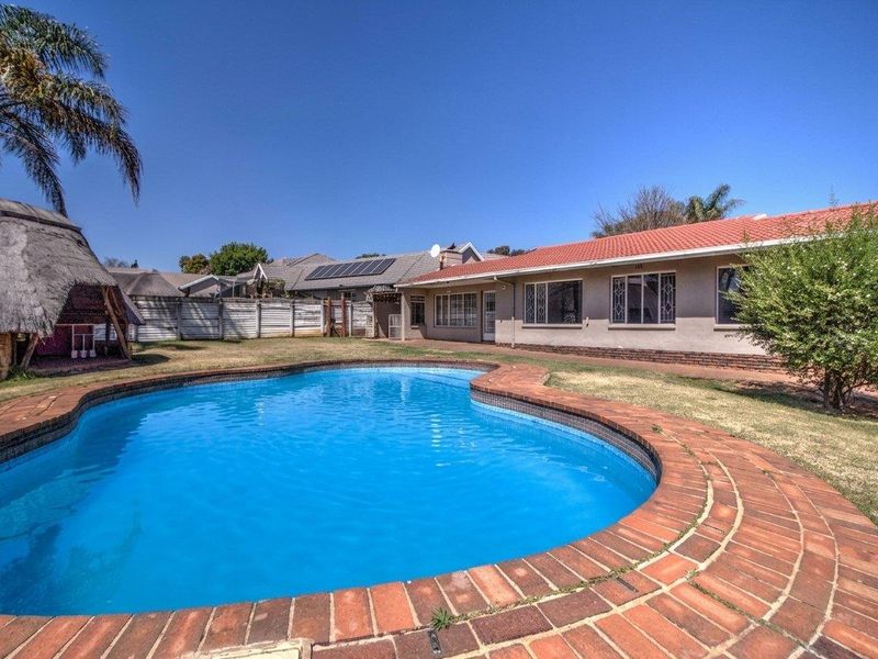 Rear garden featuring a swimming pool with brick coping, a thatched lapa, and a single-story house with a tiled roof.