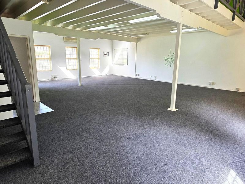 Spacious open-plan office area featuring a mezzanine level, staircase, exposed ceiling beams, and carpeted flooring, providing a versatile workspace.