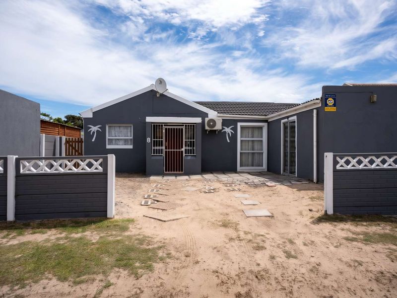 Front elevation of a single-story house featuring a dark grey facade, security gate, air conditioning unit, and a sandy front yard with paved walkway.