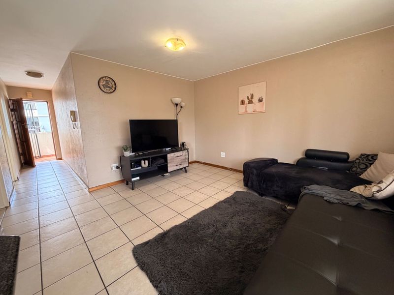 A living room area featuring neutral wall tones and tiled flooring, with an adjacent hallway leading to the entrance.