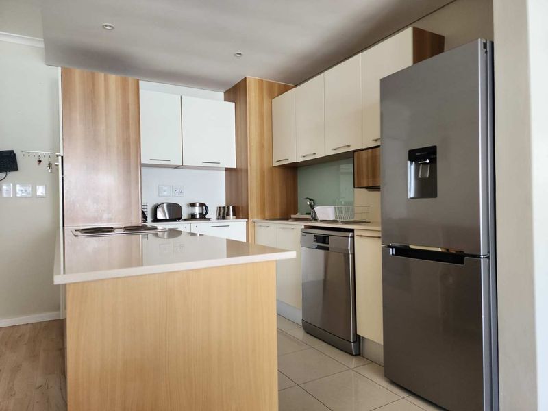 Modern kitchen featuring light wood and white cabinetry, stone countertops, integrated hob, and stainless steel appliances, including a dishwasher and refrigerator.