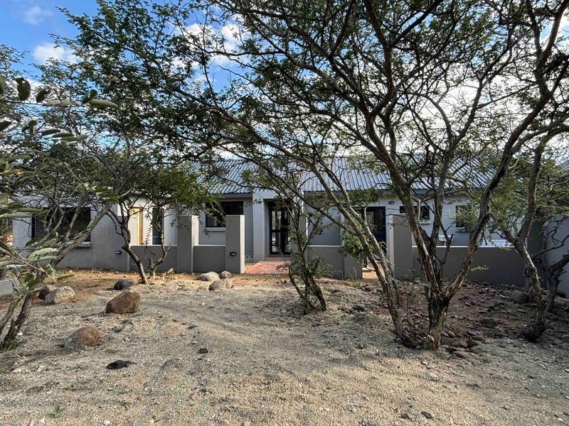 Front view of a single-story house featuring a corrugated metal roof, masonry boundary walls, and a paved entrance path surrounded by mature trees.