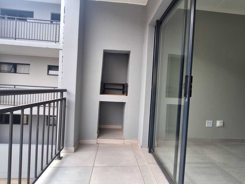 A clean, modern balcony featuring tiled flooring, a built-in braai, and a large sliding glass door providing access to the interior living space.