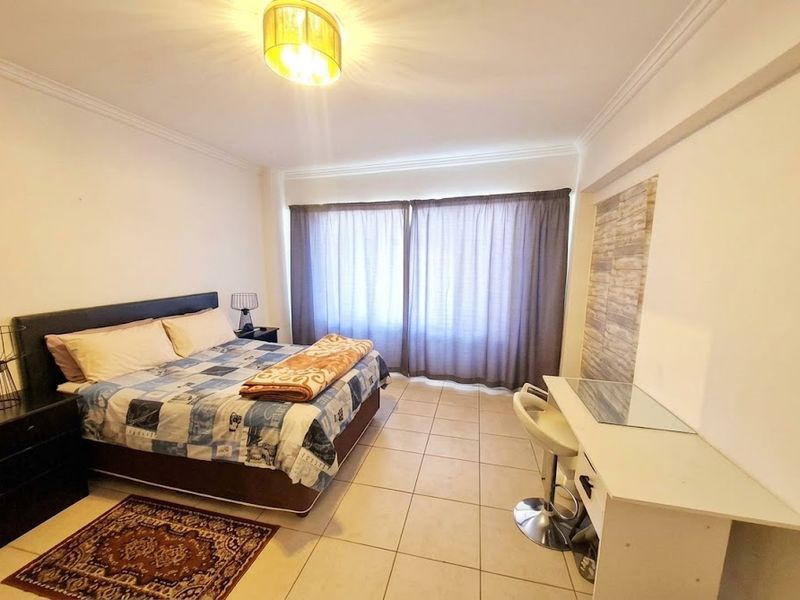 A spacious bedroom featuring tiled flooring, ceiling cornice detailing, and large windows with curtains, providing a functional and well-lit living space.