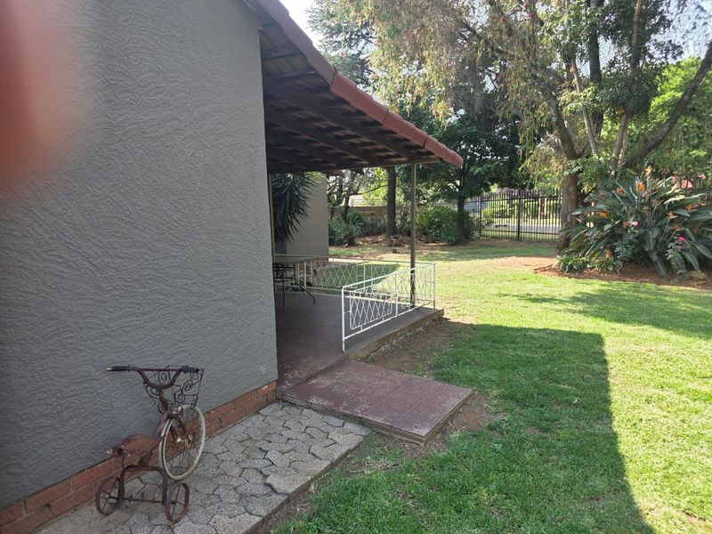 A covered patio area featuring tiled flooring and metal railings, overlooking a spacious garden with mature trees.