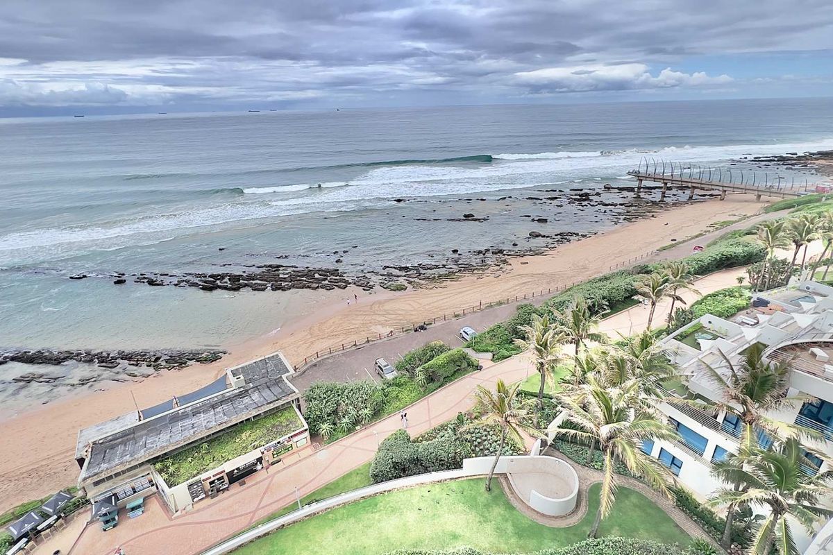 Elevated aerial view overlooking the coastline, beach, and surrounding apartment complex grounds.