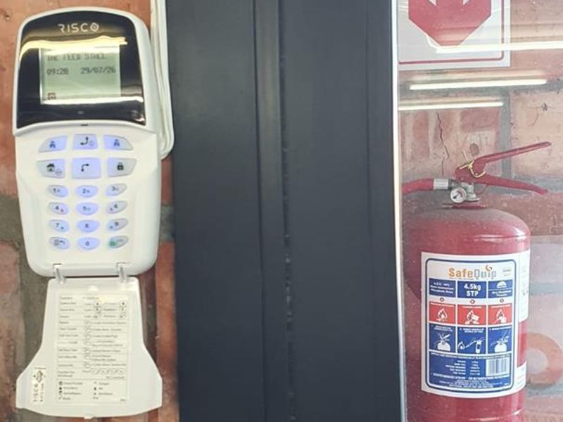 A utility area featuring an exposed brick wall, a wall-mounted security alarm keypad, and a fire extinguisher with safety signage.