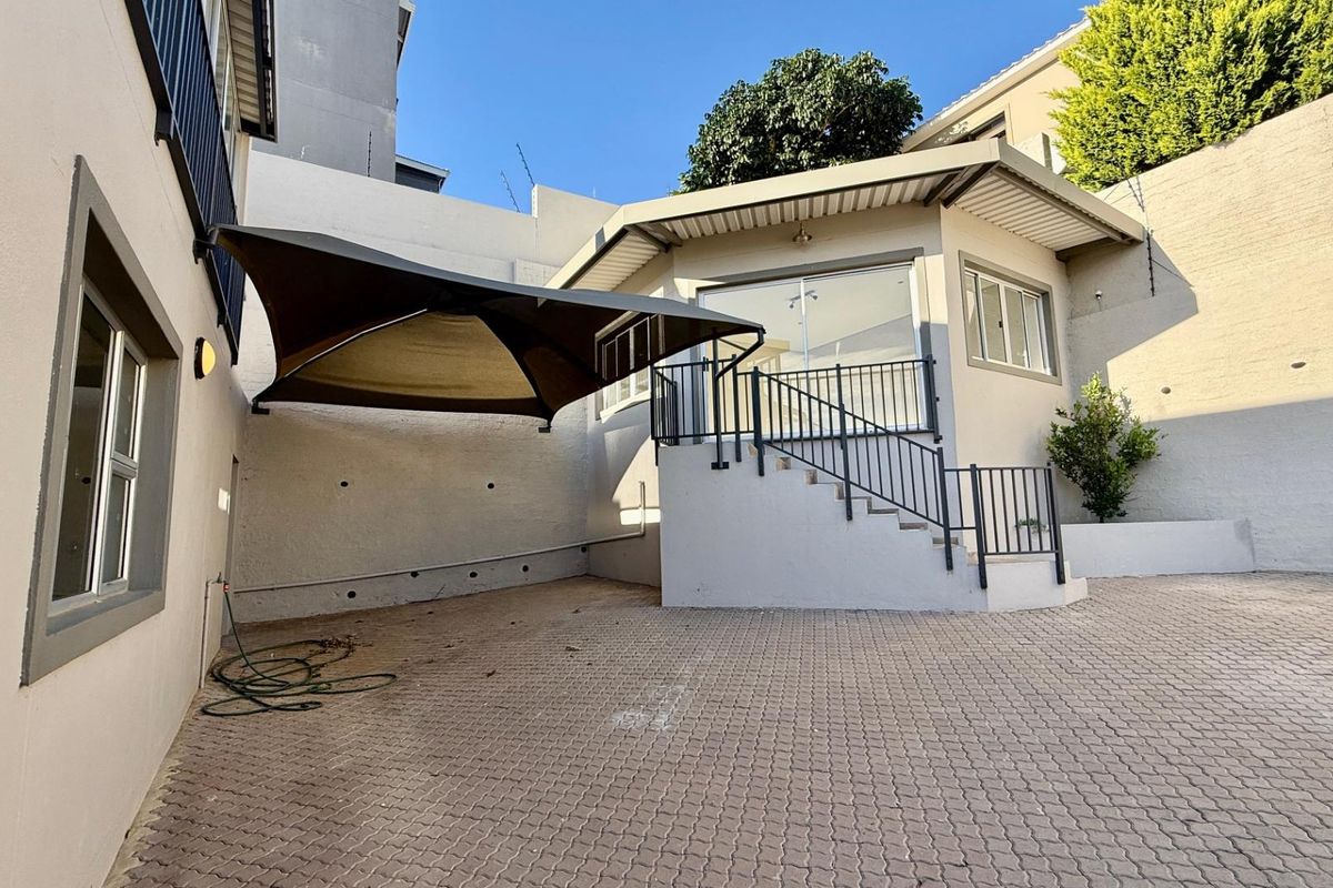 A paved rear courtyard area featuring a raised patio with metal railings, a shade sail, and exterior building access.