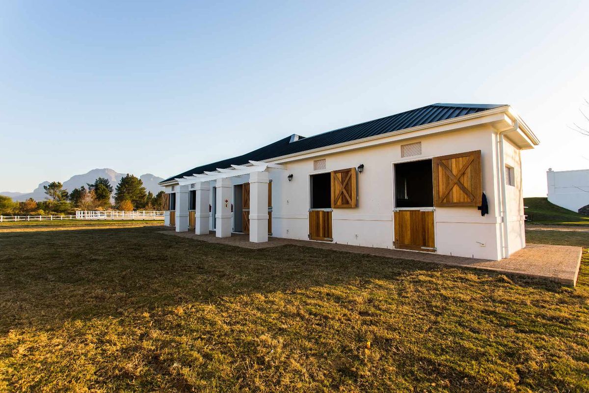 A well-maintained stable block featuring multiple individual stalls with wooden doors, a covered pergola walkway, and a metal roof set in a rural landscape.