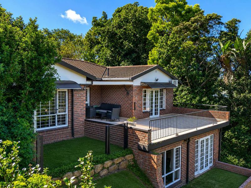 Rear view of a brick townhouse featuring a raised deck with metal railings, artificial lawn, and mature garden surroundings.