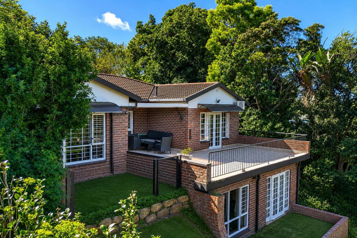 Rear view of a brick townhouse featuring a raised deck with metal railings, artificial lawn, and mature garden surroundings.