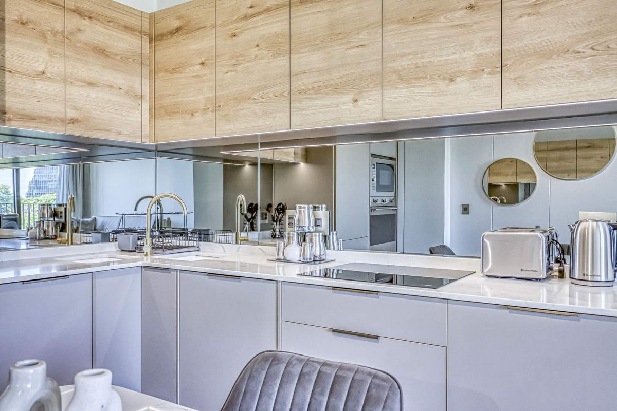 Modern kitchen featuring light wood-grain upper cabinetry, sleek grey lower cabinets, stone countertops, a mirrored backsplash, and a built-in induction cooktop.